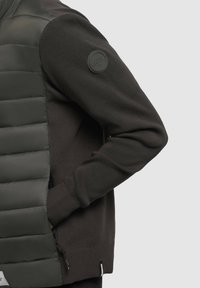 Mørkegrøn hybridjakke med polstret front og strikkede ærmer. Har en lynlås og et logo-patch på venstre ærme. Teksturerede materialer.