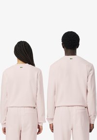 Felpa rosa chiaro con maniche lunghe, polsini a costine e un logo verde sul retro. Il tessuto appare liscio e morbido.