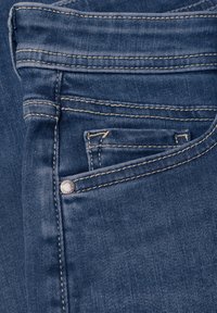 Gros plan sur un jean en denim bleu montrant la poche avant, la petite poche à monnaie, la boucle de ceinture et les détails de couture beige.