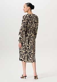 Zwart en beige patroonjurk met lange mouwen en een tailleband. Heeft een geometrisch, abstract ontwerp en een lengte tot halverwege de kuit.