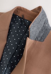Manteau marron avec col intérieur gris, doublure noire à pois blancs et une chaîne noire pour accrocher à l'intérieur de l'encolure.