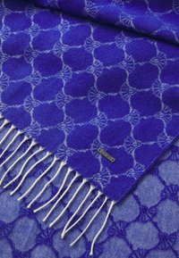 Sciarpa blu a motivo con frange bianche, caratterizzata da un design geometrico e un'etichetta in metallo. Realizzata in tessuto morbido e texture.