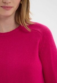 Pull en tricot fuchsia avec un col rond, une finition texturée et des manches raglan. Le tissu est doux, présentant un motif lisse et régulier.