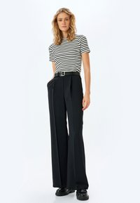 Pantalones de pierna ancha negros con pliegues frontales, combinados con una camisa de rayas en blanco y negro de manga corta y un cinturón negro. Zapatos negros gruesos.