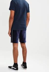 Granatowe welurowe szorty z kontrastującymi paskami po bokach, noszone z dopasowanym granatowym t-shirtem i czarnymi sneakersami. Gładka tekstura, długość do kolan.