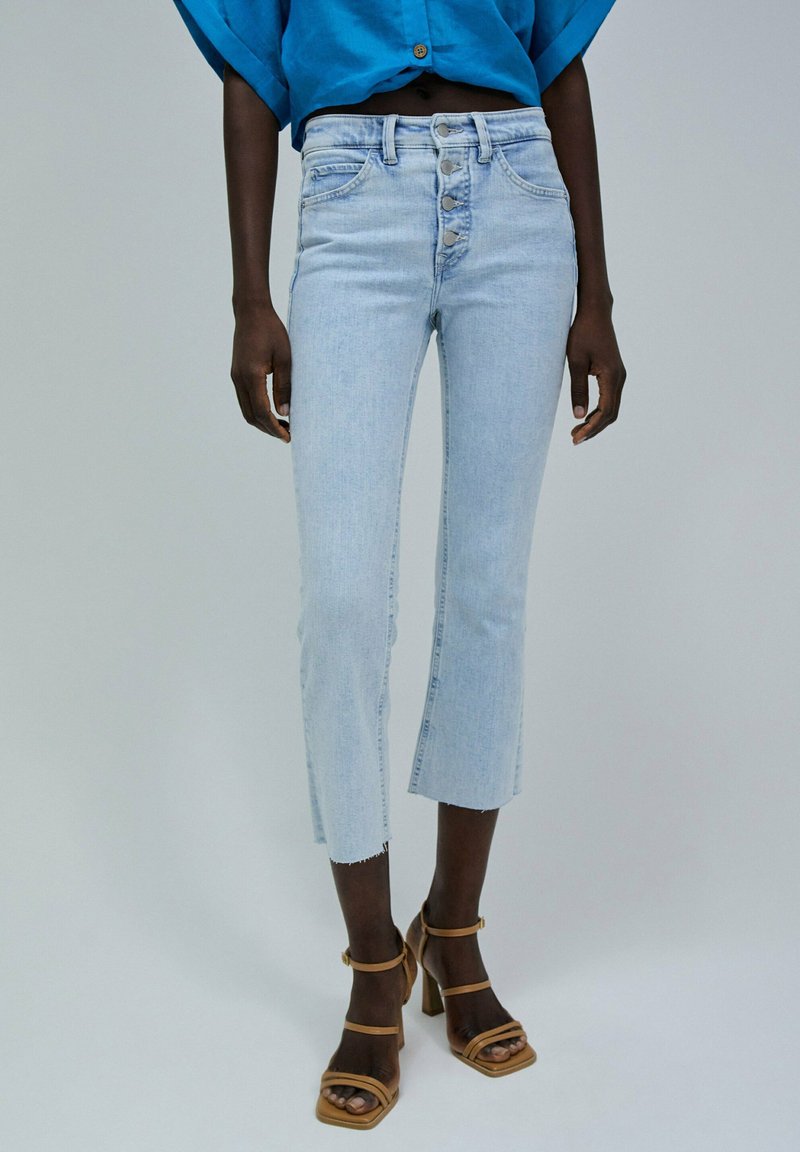 Jeans de tiro alto en azul claro con pernera acampanada y cropped; presentan un cierre de cinco botones y un dobladillo sutilmente deshilachado; combinados con tacones de bloque marrones.