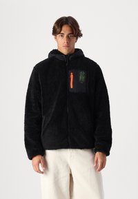 Giacca in fleece nera con zip frontale e cappuccio. Presenta una tasca nera a contrasto con zip arancione, logo verde e tessuto testurizzato.