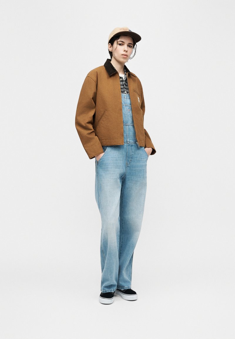 Person wearing a brown jacket over plaid shirt and light blue denim overalls, beige cap, and black slip-on shoes, standing with hands in pockets.