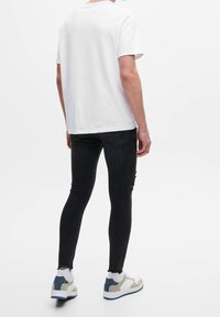 Persona vestida con una camiseta blanca sencilla, unos jeans ajustados negros con rotos, calcetines blancos y zapatillas blancas con detalles en azul y beige, de pie de espaldas.
