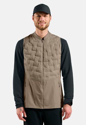 Mann trägt eine gesteppte beige Weste über einem schwarzen Langarmshirt und einer schwarzen Mütze, lächelt, steht vor einem schlichten weißen Hintergrund.