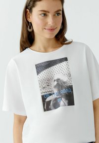 Witte t-shirt met korte mouwen, voorzien van een fotografische print met zwart-wit afbeeldingen, blauwe tekst en een textuurpatroon accent.