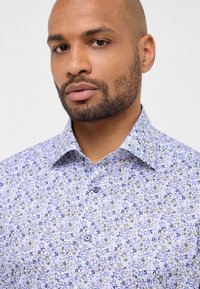 Camicia a fantasia floreale in azzurro chiaro, con piccoli fiori viola e verdi. Il collo button-down e i bottoni scuri aggiungono dettagli.