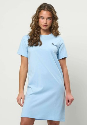 Młoda kobieta o falujących brązowych włosach, ubrana w jasnoniebieską, krótkorękawową, casualową sukienkę z małym haftowanym logo na piersi.