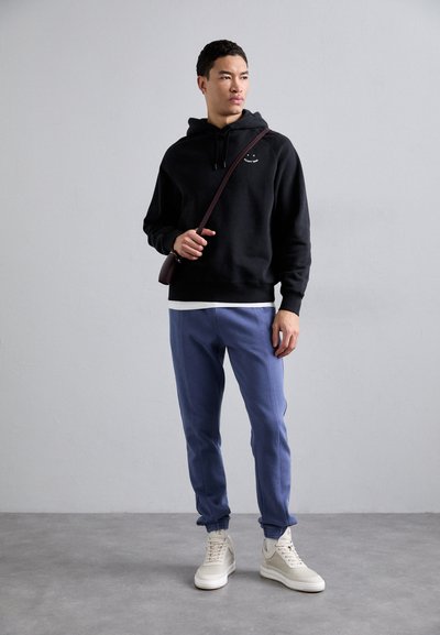 Fekete pamut kapucnis pulóver hímzett részlettel, világoskék szűkített melegítőnadrággal és fehér magas szárú sneakers-szel párosítva. A modell egy kis táskát tart.