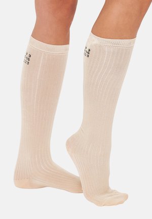 Chaussettes beiges montantes avec une texture côtelée et un renfort au niveau des orteils. Présente des chiffres noirs imprimés sur le bandeau supérieur. Tissu doux et extensible.