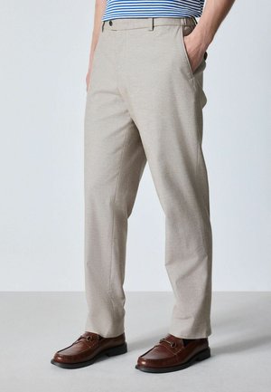 Mann trägt beige Hose, braune Leder-Loafers und ein blau-weiß gestreiftes Hemd mit Händen in den Taschen.