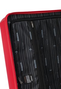 Valigia rigida rossa con texture rigata, interno nero con motivi stampati e una tasca con zip, include un dettaglio con etichetta.