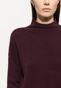 Frau, die einen dunkelvioletten Rollkragenpullover und einen kleinen goldenen Creolenohrring trägt, von den Schultern bis zum Kinn vor einem neutralen Hintergrund gezeigt.