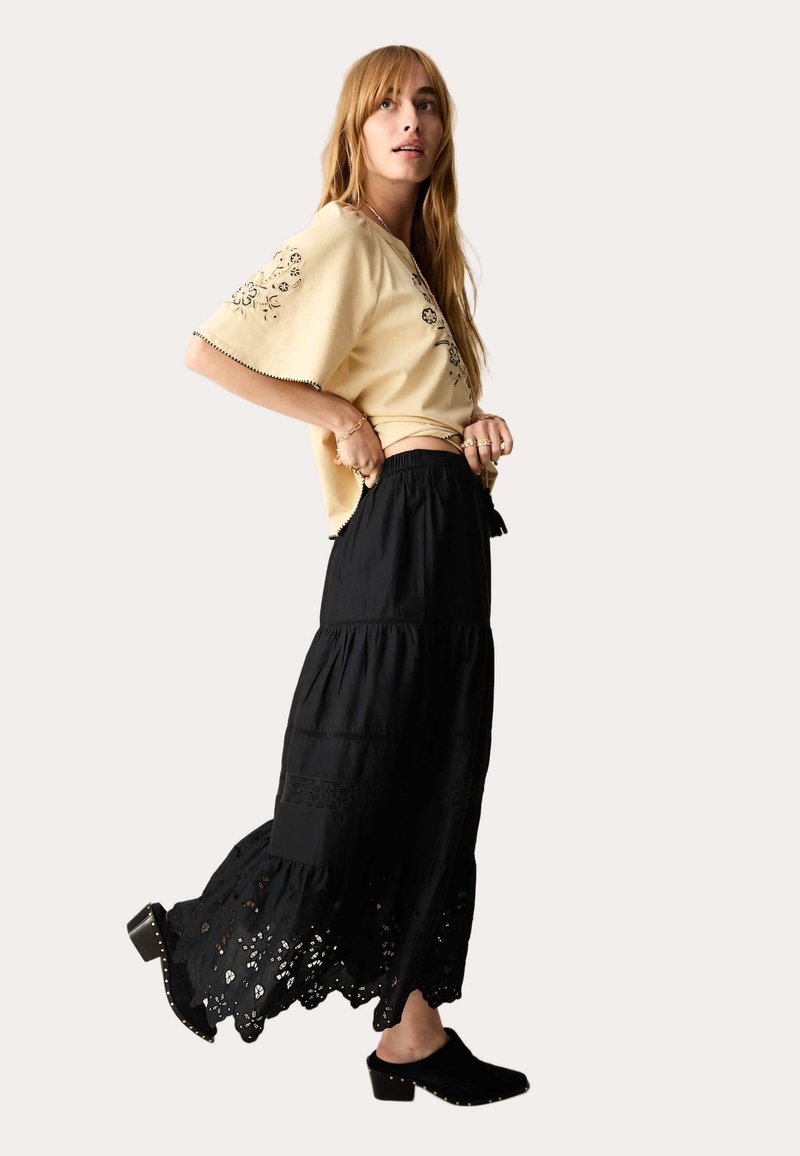Donna che indossa una blusa ricamata beige e una lunga gonna nera a strati con dettagli in pizzo ritagliato, mentre regola la sua gonna guardando verso l'alto.
