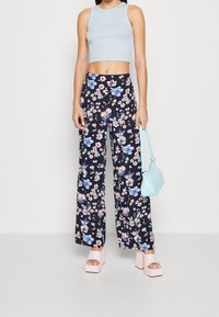 Femme portant un crop top bleu clair, un pantalon large à fleurs noir, des sandales à plateforme roses, tenant un sac à main bleu clair, sur fond blanc.