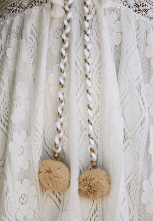 White lace garment featuring floral patterns, with braided tie accents in white, brown, and gold, topped with two round beige pom-poms.