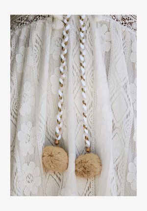 Indumento in pizzo bianco con motivi floreali, decorato con legature intrecciate in bianco, marrone e oro, completato da due pom-pom rotondi beige.