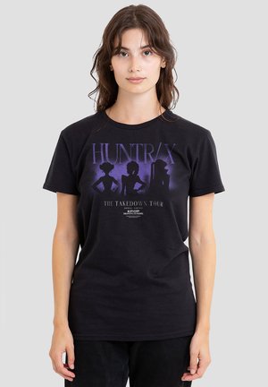 Young woman wearing a black Huntrax "The Takedown Tour" graphic t-shirt featuring three purple silhouettes of female figures.
