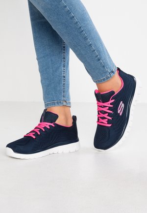 Person iført blå mesh-sneakers med lyserøde snørebånd og hvide såler, kombineret med lyseblå tætsiddende jeans, stående på et hvidt gulv.