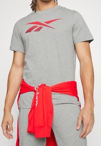 Muž v šedém tričku Reebok s červeným logem, šedých kalhotách a červené mikině svázané kolem pasu.