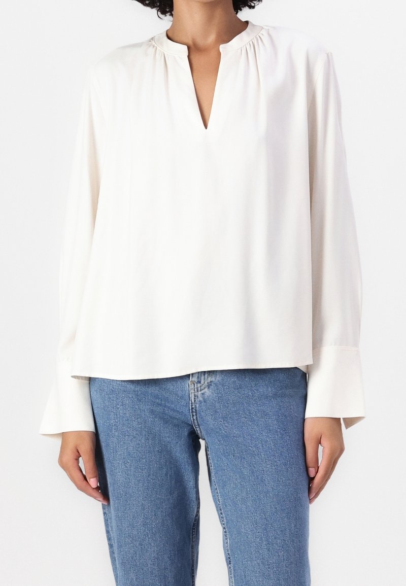 Blouse blanche à manches longues avec une coupe décontractée, un col en V, un détail froncé au niveau du col et des poignets évasés, associée à un jean en denim bleu.
