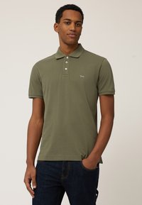 Polo de color verde oliva en tejido suave, con cuello de tres botones y mangas cortas. Acento de logo pequeño en el lado izquierdo del pecho.