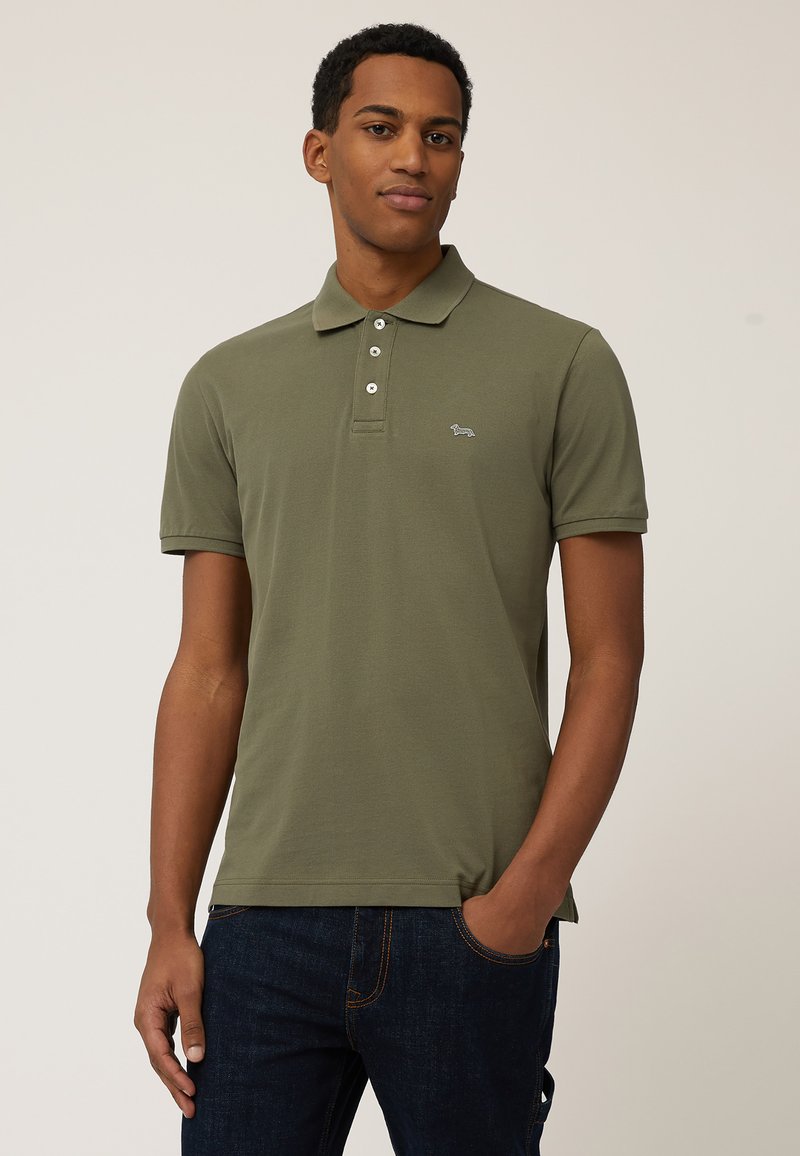 Polo de color verde oliva en tejido suave, con cuello de tres botones y mangas cortas. Acento de logo pequeño en el lado izquierdo del pecho.