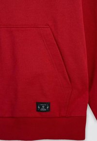 Czerwona bluza z kapturem wykonana z miękkiej bawełny, posiada kangurzą kieszeń, ściągacze przy mankietach i dolnym końcu, z czarną metką na dole.