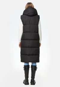 Svart vadderad väst med huva, med horisontella quiltade sektioner. Modellen bär blå jeans och knähöga svarta stövlar med spännen.