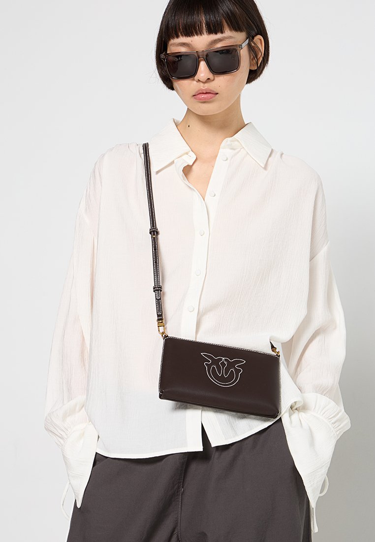 Giovane donna con capelli corti, che indossa grandi occhiali da sole neri, una camicia bianca sbottonata, pantaloni scuri e una borsa a tracolla nera con ricami bianchi.