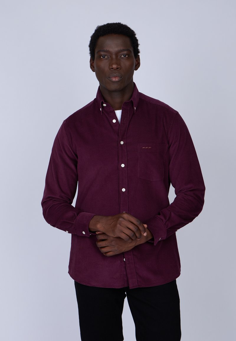 Chemise en velours côtelé bordeaux avec une poche poitrine, boutons blancs et texture lisse, portée avec un pantalon noir sur fond uni.