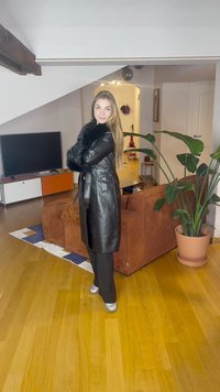 Cappotto lungo in pelle nera con collo in pelliccia sintetica, cintura in vita e design aderente. In piedi su pavimento in legno con piante e mobili sullo sfondo.