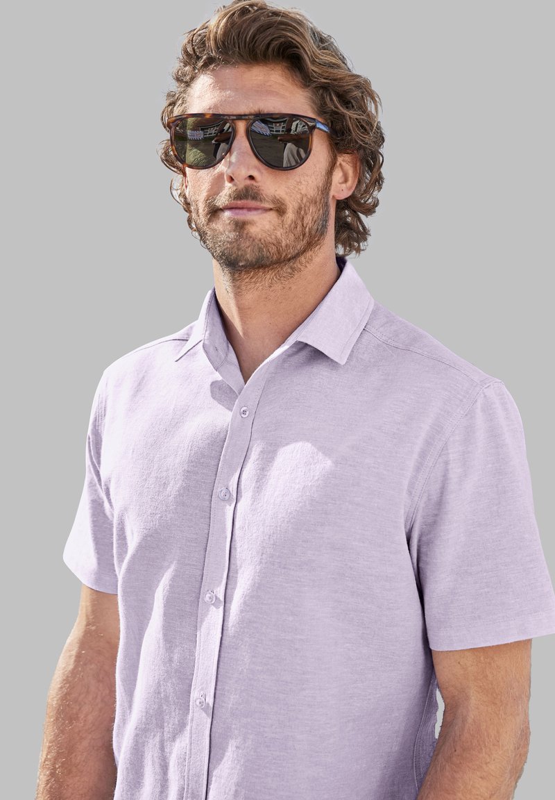 Korte mouwen, lichtpaarse blouse met knoopjes, gemaakt van textuurstof. De drager draagt ook reflecterende zonnebril met een bruine montuur.