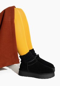 Botines de ante negro con suela gruesa, combinados con medias amarillas y una falda marrón. Presentan un detalle de calcetín negro texturizado y acanalado.