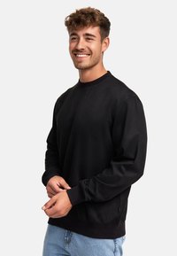 Sudadera negra de manga larga y cuello redondo, con una textura suave y dobladillo acanalado. Combinada con jeans de color azul claro.
