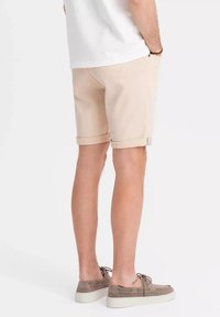 Shorts beiges avec un ourlet retroussé, fabriqués à partir d'un tissu respirant, associés à des chaussures à lacets marron, vus de l'arrière. Design et texture simples.