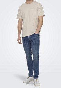 Camiseta de rayas beige con un bolsillo en el pecho, combinada con jeans de denim azul oscuro. El modelo lleva zapatillas blancas y está de pie frente a un fondo claro.