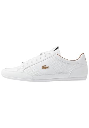 White low-top sneaker with embossed text pattern, brown crocodile logo on side, white laces, and a slim white sole.
