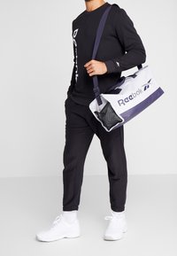 Bolsa de viaje Reebok en blanco y marino, con un bolsillo de malla, dos asas y una correa para el hombro ajustable, diseñada para un uso versátil.