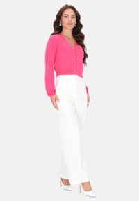 Růžový kardigan s dlouhými rukávy a knoflíky, kombinovaný s bílými kalhotami s vysokým pasem. Modelka má na sobě bílé lodičky. Jednoduchý design, hladká textura.