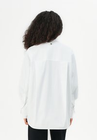 Witte blouse met lange mouwen en een comfortabele pasvorm, voorzien van een plooitje aan de achterzijde en een decoratieve knoopdetail op de kraag. Soepele stof.