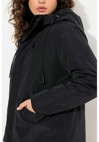 Zwarte, waterdichte jas met een capuchon, ritssluiting aan de voorkant en een trekkoord. Opvallende kenmerken zijn een borstzak en een merkpatch op de mouw.