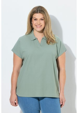 Uśmiechnięta kobieta z blond włosami w jasnorzęowej, krótkim rękawem z kołnierzykiem i niebieskich dżinsach, stojąca na białym tle.