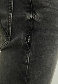 Jeans de mezclilla negros de corte slim con una textura ligeramente desgastada. Presenta un bolsillo lateral con una etiqueta de logo visible y detalles de costura en contraste.