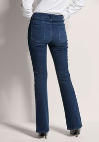 Dark blue denim flared jeans with a high waistband, featuring two back pockets and a smooth texture, paired with black heeled shoes.